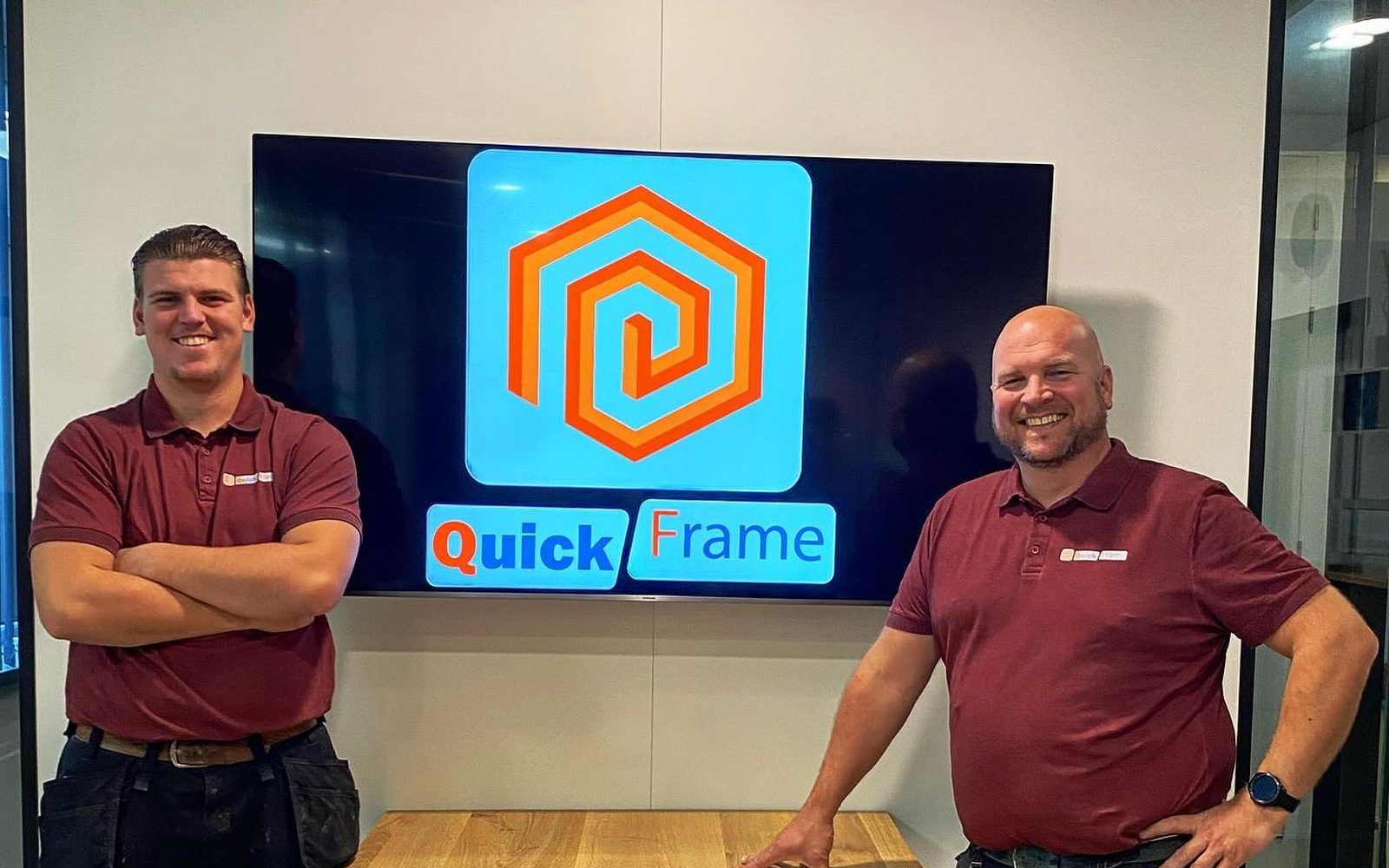Quint en Daniel Vaalburg in werkkleding van Quickframe, voor een scherm met het logo